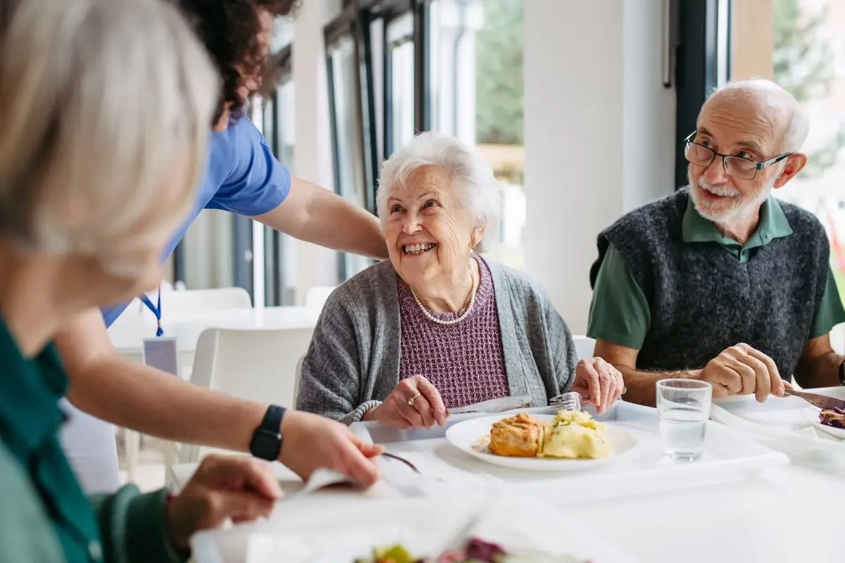 A carer in a blue uniform serving lunch to smiling older residents at a table in a bright care home dining room