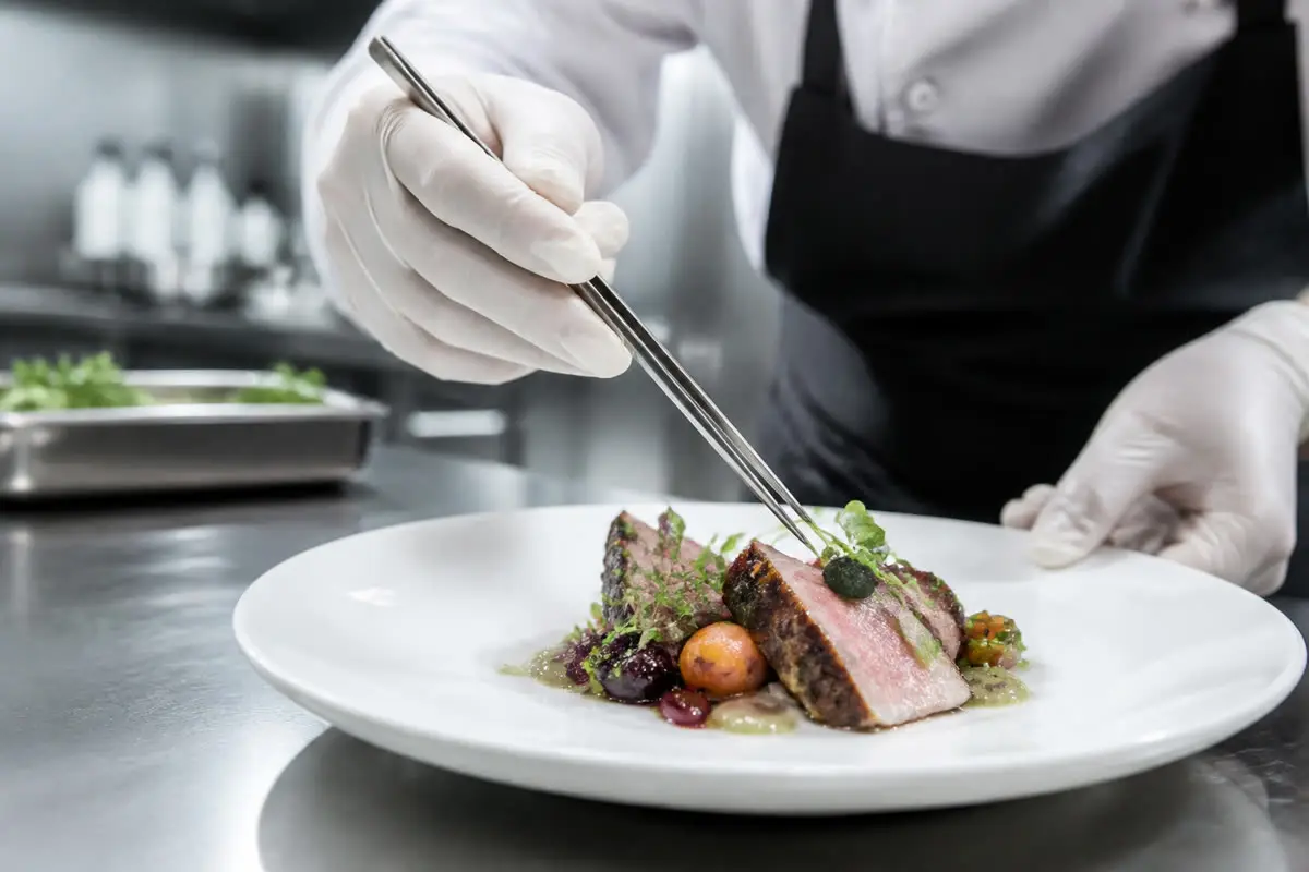 Chef wearing disposable gloves plating a dish on a stainless steel pass in a professional kitchen